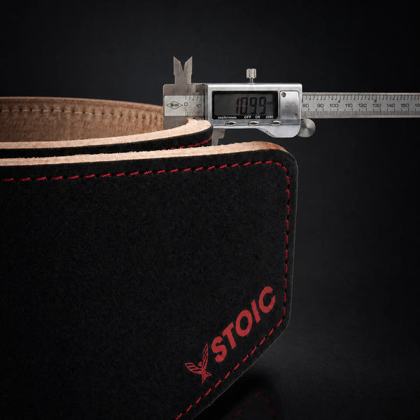 Stoic 10mm single-prong weight lifting belt in Bloodline red and black, a digital caliper measuring the belt's thickness, its display reading 10.99 mm.