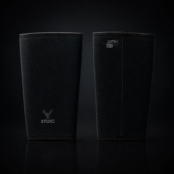 Stoic 7mm knee sleeves for weightlifting in Black Label, the pair standing side by side, one showing the phoenix logo on its front and the other showing the woven size and care label.