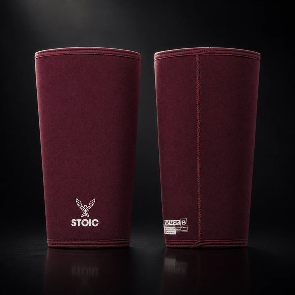 Stoic 7mm knee sleeves for weightlifting in maroon, the pair standing side by side, one showing the phoenix logo on its front and the other showing the woven size and care label.