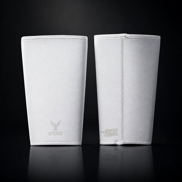 Stoic 7mm knee sleeves for weightlifting in white, the pair standing side by side, one showing the phoenix logo on its front and the other showing the woven size and care label.