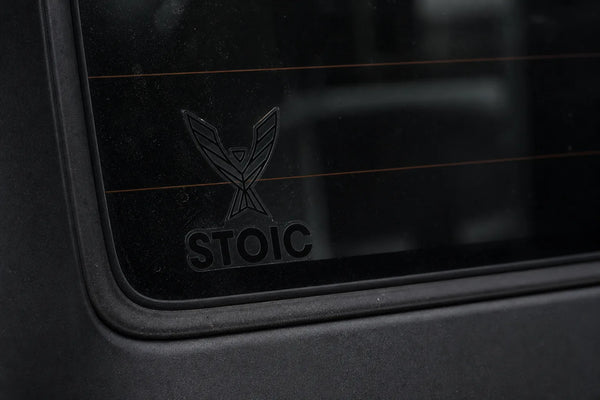 Stoic clear vinyl decal, 2.75 inch, applied to a vehicle rear window - the glass, defroster line and view through the transparent film are visible, showing it has no white backing